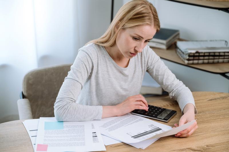 Une femme concentrée travaille, en utilisant une calculatrice et consultant plusieurs documents étalés devant elle.